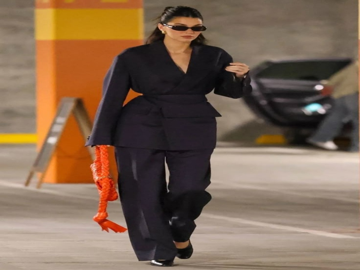 A model showcasing a tailored blazer and wide-leg trousers, walking confidently in a stylish setting with a vibrant handbag.