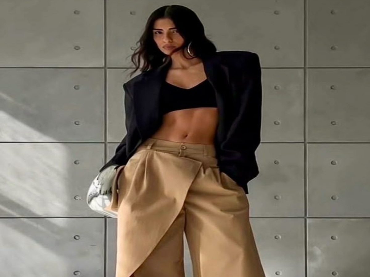 Model in a black blazer over a cropped top, paired with high-waisted pleated trousers, posing against a modern concrete backdrop.