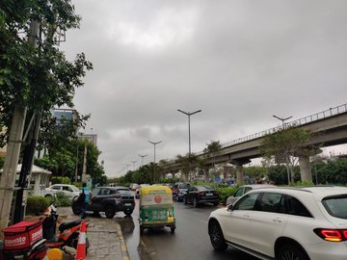 Urban road with cars, bikes, and auto rickshaws under cloudy skies near a metro line in the city.