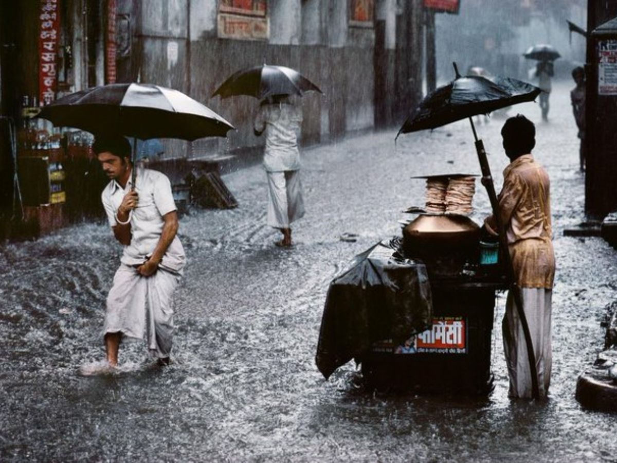 Humans taking walks and running in heavy monsoon rain on a flooded avenue in India, with umbrellas and a roadside meals stall seen.