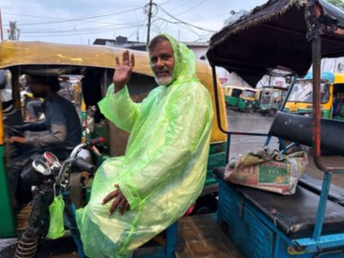 A rickshaw motive force in a inexperienced raincoat smiles and waves all through monsoon, with car-rickshaws inside the historical past.