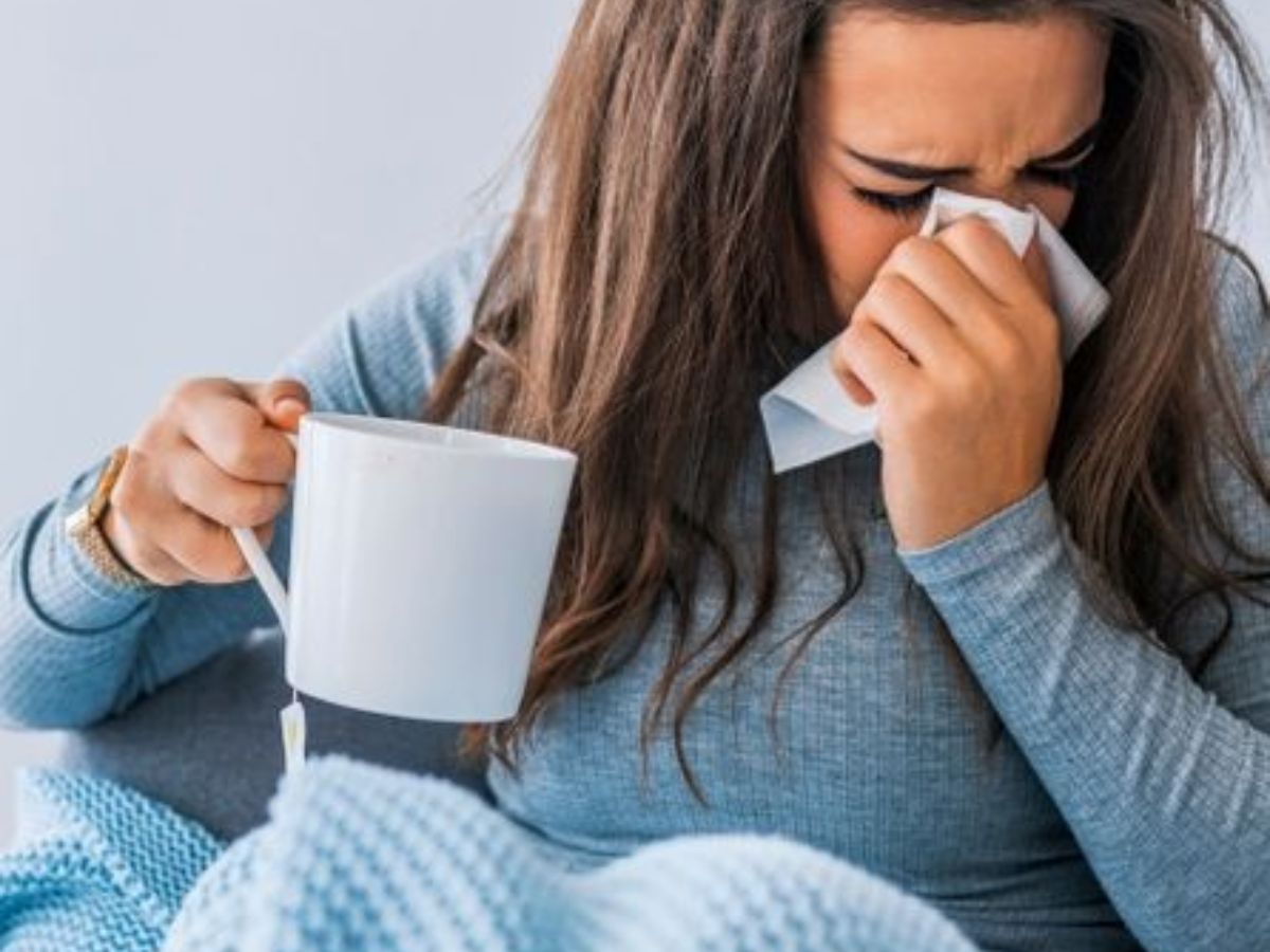 A sick woman in a sweater holding a cup and wiping her nose with a tissue, showing signs of cold and fatigue.