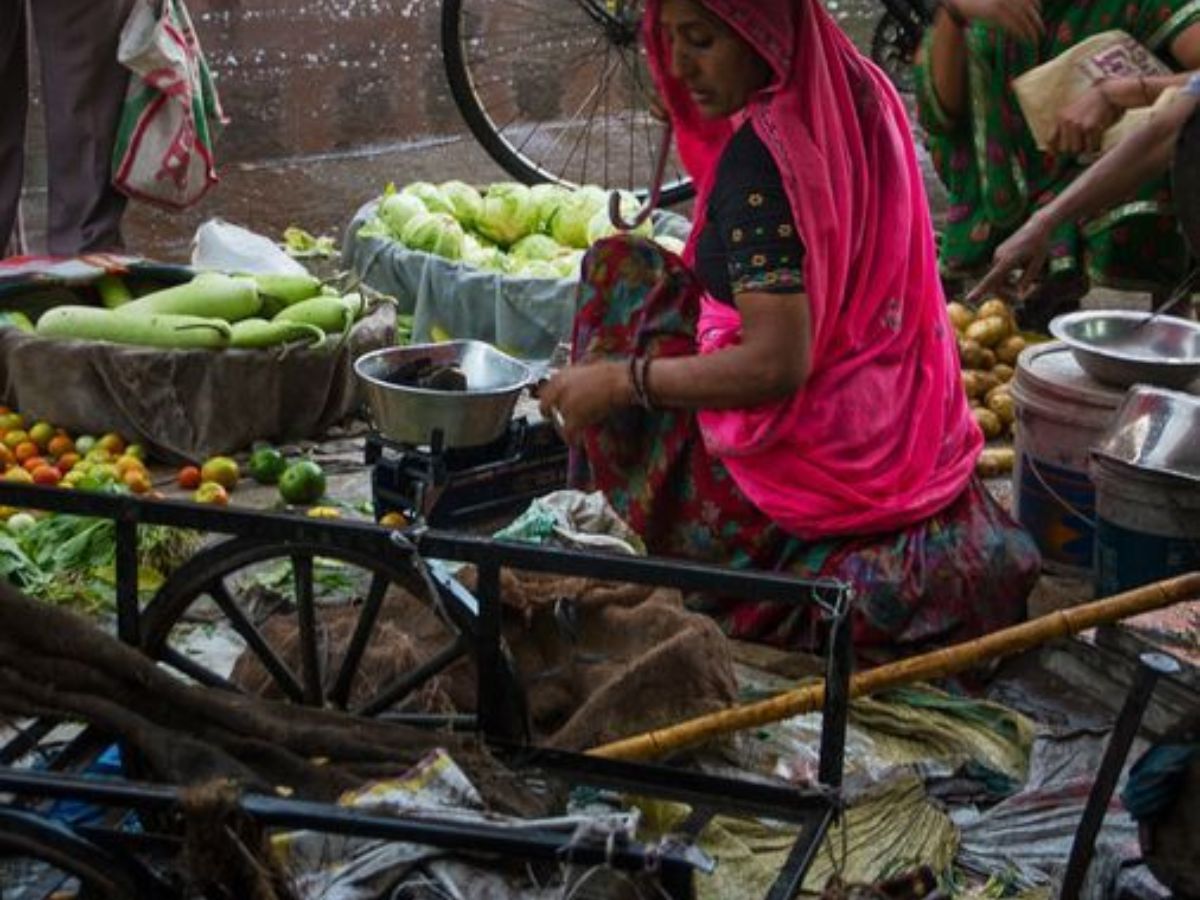 A woman selling vegetables on a damp roadside market in monsoon, surrounded by wet sacks and produce.