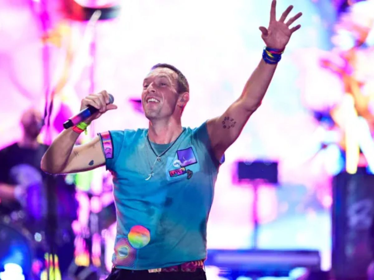 Chris Martin performing energetically onstage with colorful lights in the background