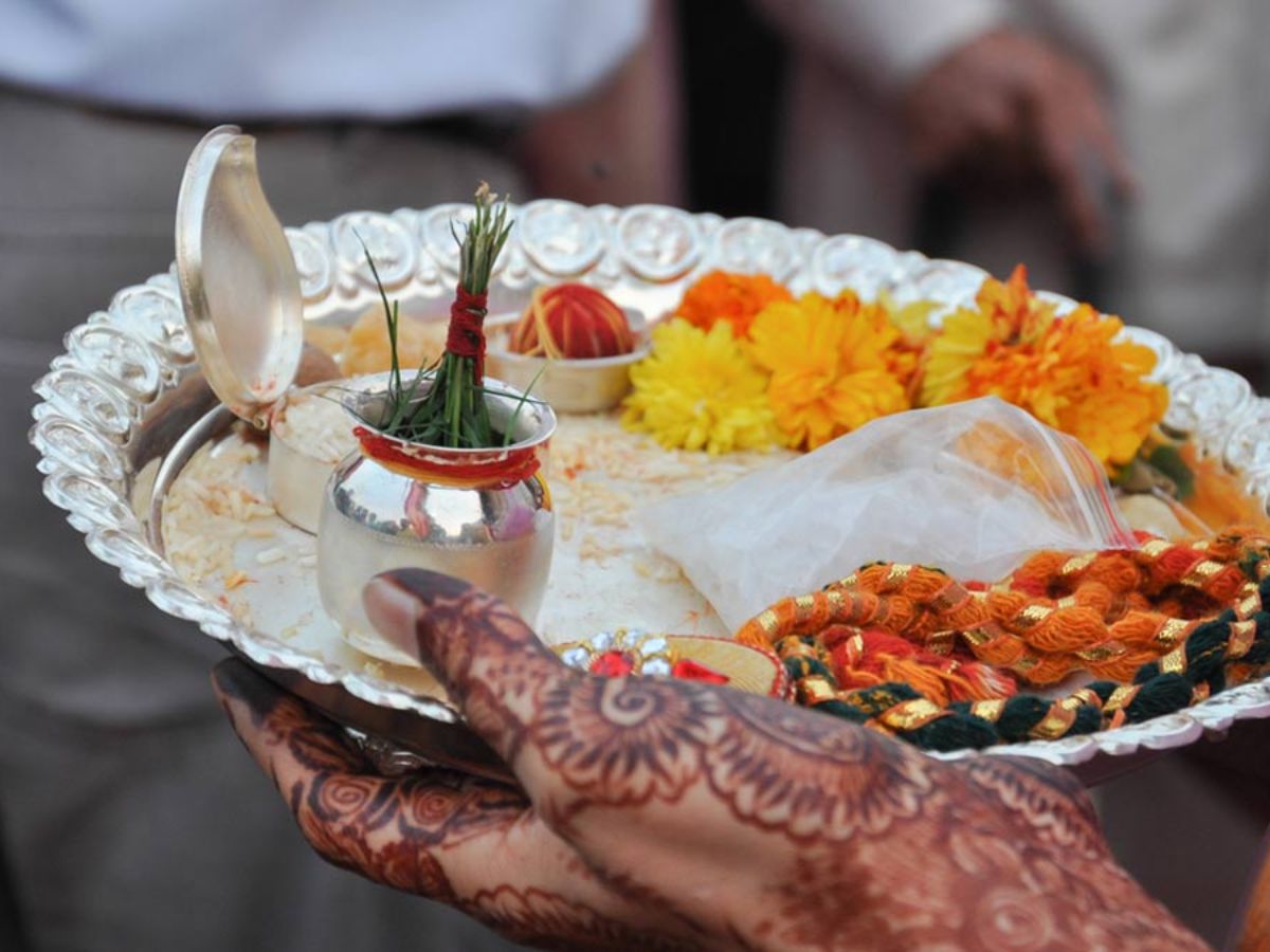 A decorated puja thali held by a woman with mehendi on her hands, containing kalash, flowers, rice, sacred thread, and other items used during Shivratri fasting rituals