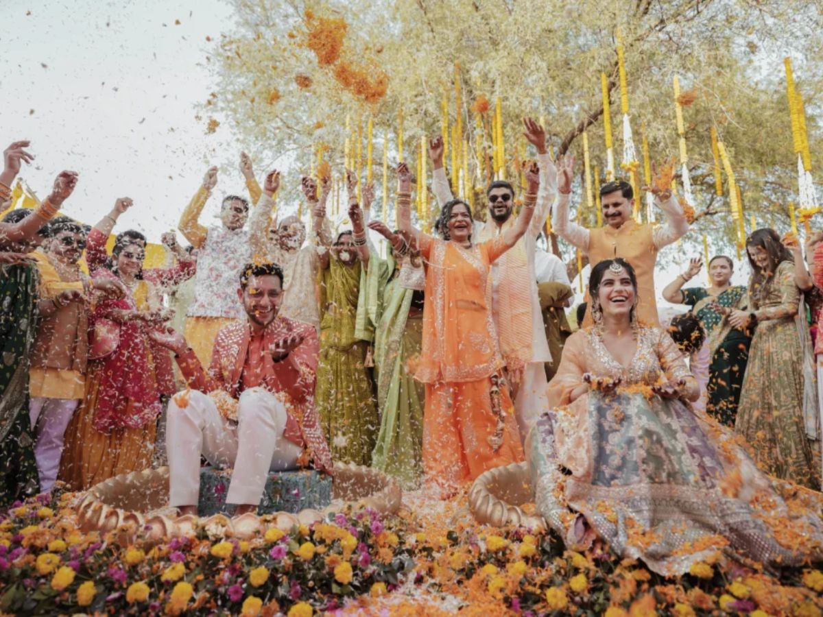 Bride and groom seated joyfully during a vibrant Haldi or pre-wedding ceremony, surrounded by family and friends showering them with flower petals, dressed in colorful traditional outfits and celebrating outdoors under floral decor
