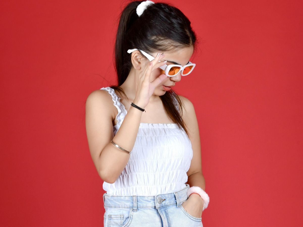 A young woman with a high ponytail wears a white smocked crop top, light-wash jeans, and white-framed sunglasses with orange lenses. She is adjusting her sunglasses with her left hand, with her right hand in her pocket. The background is a solid red color.