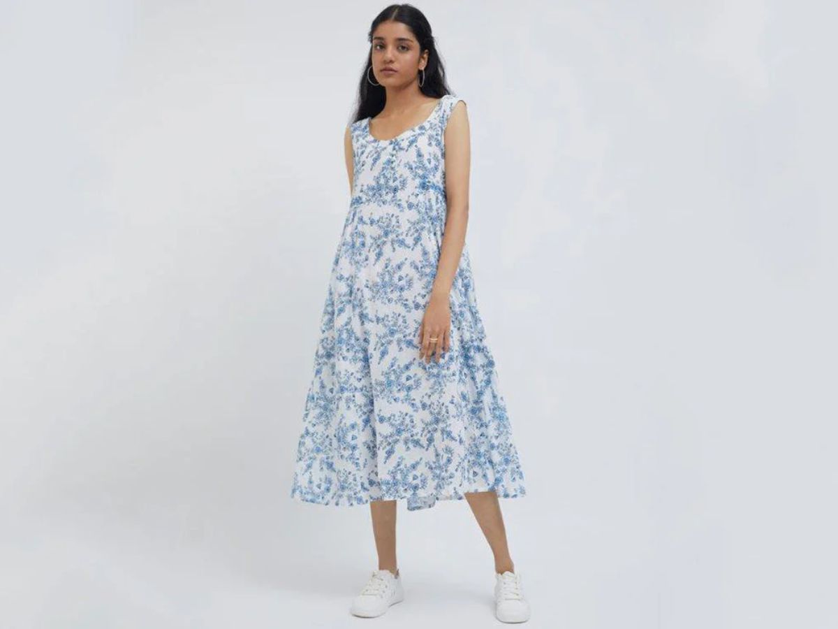A full-length shot of a young woman standing on a white background, wearing a white sleeveless A-line dress with a light blue floral pattern and white sneakers. She has on simple hoop earrings and is looking directly at the camera.
