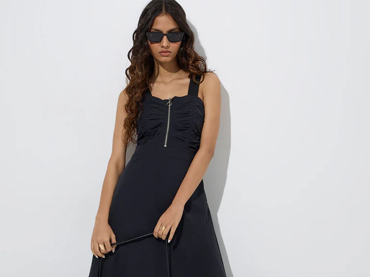 A medium close-up shot of a woman with long, wavy hair, wearing a black sleeveless dress with a zippered front and a ruched bust. She is also wearing black sunglasses and multiple rings. She is holding a thin-strapped black bag and standing in front of a white wall.