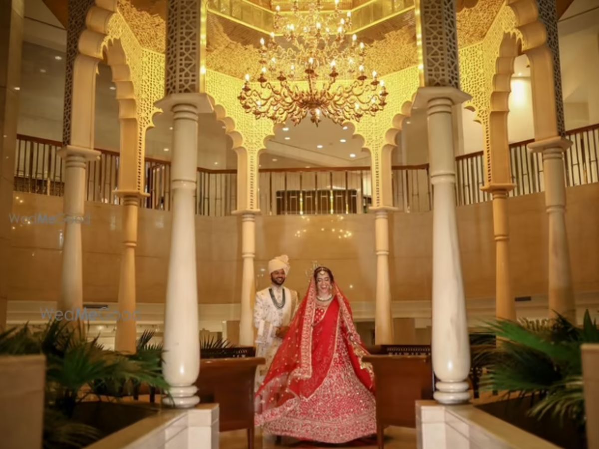 A bride and groom in traditional Indian wedding attire stand smiling in a lavish, ornate lobby with grand pillars, detailed arches, and a large chandelier.
