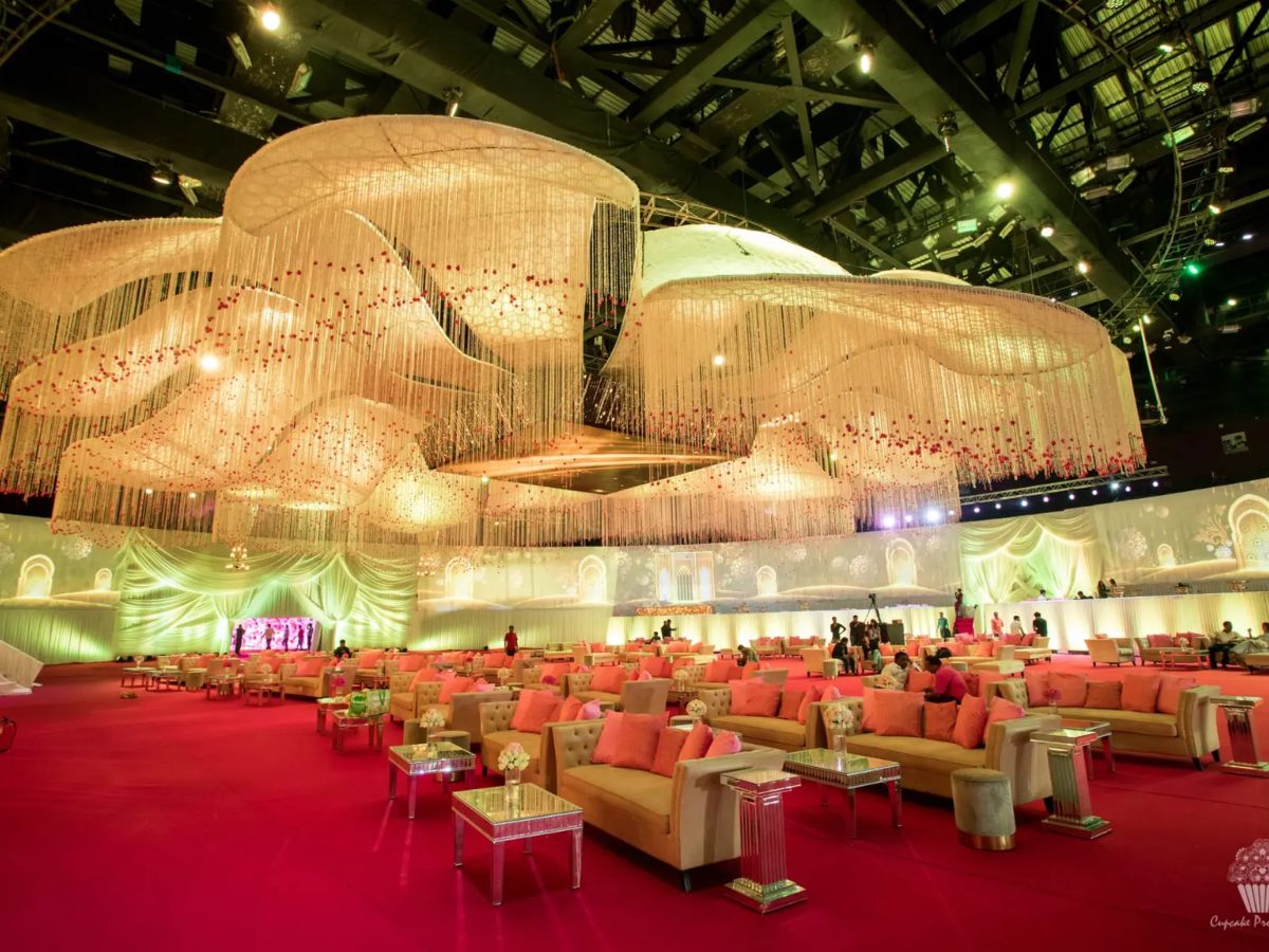 A large, luxurious indoor event space with a red carpet and rows of plush beige sofas with orange pillows. The ceiling is extravagantly decorated with massive, curved white floral installations and chandeliers, creating a grand and opulent atmosphere.
