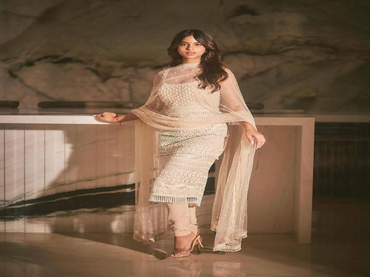 A person wearing an elegant cream traditional outfit with intricate embroidery and a sheer dupatta, posing in a sophisticated setting.